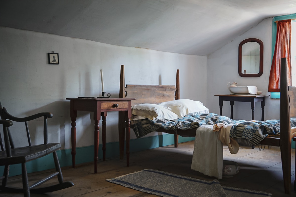 small bedroom with iron bedstead, lace curtains, and a candle on the dresser